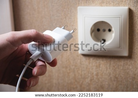 Hand turns on, turns off charger in electrical outlet on wall. White charger for phone in woman's hand close-up