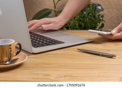 Hand Is Typing On Laptop, Second Hand Is Holding Smartphone, Pen And Cup Of Coffee On Light Wooden Table. Work From Home
