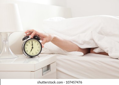 Hand under blanket reaching out for alarm clock, shallow depth of field focus on foreground