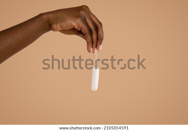 Hand of\
unrecognizable black woman holding tampon over beige studio\
background, isolated, copy space, cropped. Female period and\
intimate hygiene, menstrual products\
concept