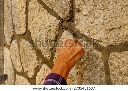 A mason’s hand uses a small tool to shape fresh mortar, carefully finishing joints in a traditional stone wall outdoors