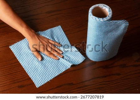 Hand using blue zigzag cleaning cloth on wooden surface, with cleaning roll beside. Household reusable and disposable wipes for kitchen and multipurpose cleaning.