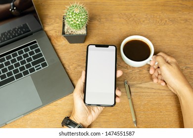 Hand using cell phone and holding coffee cup with laptop on wooden desk. - Shutterstock ID 1474551587