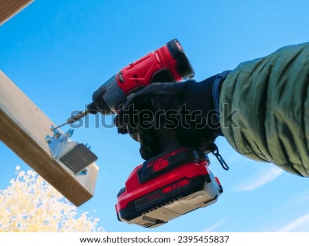 Hand using cordless impact driver fasten attach hex head exterior wood screws to galvanized joist hanger support underneath rafter beam outdoor structure, sunny sky fall foliage background, Texas. USA
