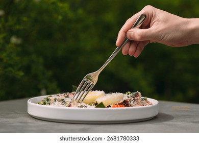Hand using a fork taking a portion of healthy food served on a white plate with green foliage in the background outdoors