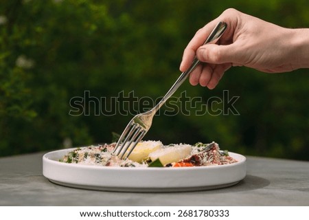 Hand using a fork taking a portion of healthy food served on a white plate with green foliage in the background outdoors