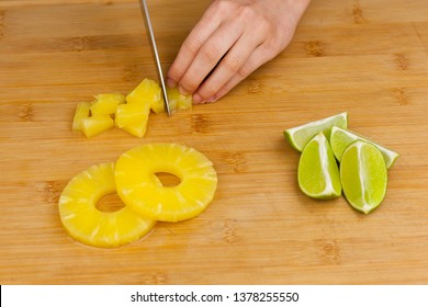 Hand using knife to cut pineapple slices and limes on a wooden cutting board background.