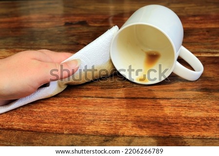 A Hand Using A Paper Towel To Clean Up A Coffee Spillage Off A Wooden Table.