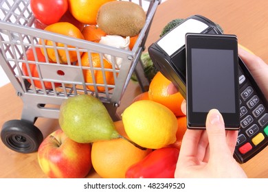 Hand using payment terminal, credit card reader with mobile phone with NFC technology and fresh fruits and vegetables with plastic shopping carts, cashless paying for shopping