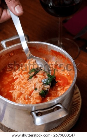 hand using a spoon to serve a portion of a hearty rice dish from a rustic metal pot. The dish, a warm, red tomato-based rice stew, is infused with vibrant green herbs.