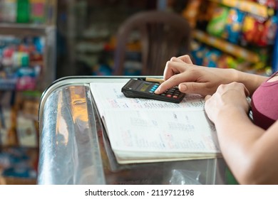 Hand View Of Latina Woman Using Her Small Calculator And Eco-friendly Cardboard Pen On A Large Notebook. Doing Calculations On Sales And Statistics In Her Grocery Store.