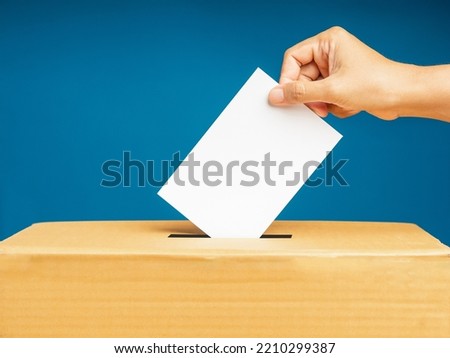 Hand voter holding ballot paper putting into the voting box at place election against a blue background. Freedom democracy concept. Close-up photo