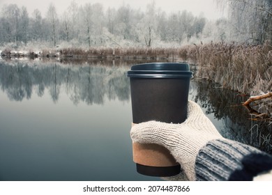 A Hand In A Warm Mitten Holds An Eco-friendly Paper Cup With Coffee On The Shore Of The Lake. Travel To Nature.