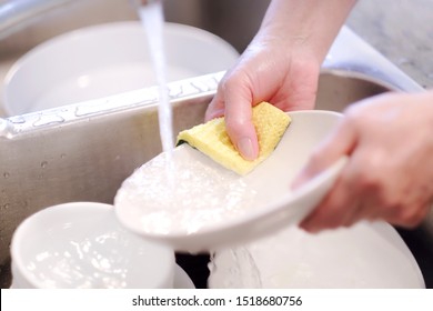 Hand Washing And Cleaning Dishes In Kitchen Sink Using Sponge Under Running Water From Faucet. Close Up And Selective Focus.