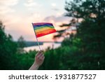 A hand waves a colorful gay pride LGBT rainbow flag at sunset on a natural landscape in summer