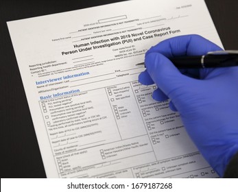 A Hand Wearing A Blue Nitrile Glove Is Shown Starting To Fill Out A Government-issued Coronavirus Covid-19 Medical Report Form.