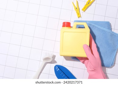 A hand wearing a pink rubber glove holds a yellow cleaning bottle, a brush, a cleaning cloth and two clothespins displayed on a checkered white tile floor. - Powered by Shutterstock