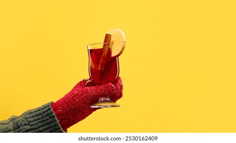 Hand wearing red knitted mitten holds glass of mulled wine, garnished with an apple slice and cinnamon stick, against warm yellow studio background. Concept of Festive winter drink, celebration. Ad - Powered by Shutterstock