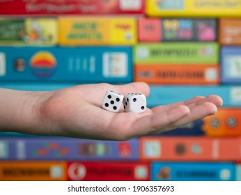 Hand With White Dice On The Blurred Background Of Colorful Board Game Boxes. Concept Of Entertainment During Pandemic Of Covid-19