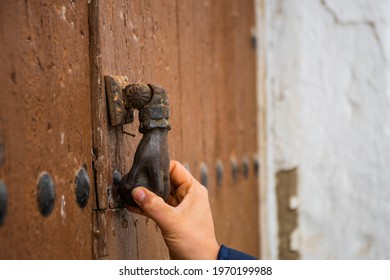 Hand Of A Woman Catching An Antique Hand Shaped Knocker With A Ball On A Wooden Door With Large Iron Nails