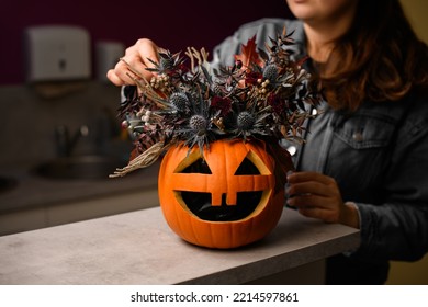 hand of woman florist gently decorate lovely autumn arrangement of thistles flowers and plants in bright orange pumpkin with carved eyes and mouth. Beautiful floral decor for halloween. Close-up - Shutterstock ID 2214597861