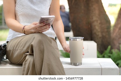 Hand of a woman grabbing reusable coffee cup, tumbler while texting message in her smart phone. Environmental friendly, eco living, No one-time use plastic, No straw, Urban leisure, Zero waste concept