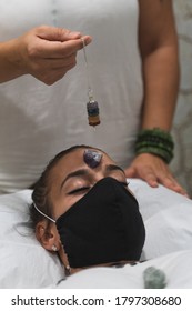 Hand Of A Woman Healer Applying Reiki And Energy Therapies Through The Use Of A Pendulum, To A Young Woman With A Medical Mask During The Coronavirus Crisis