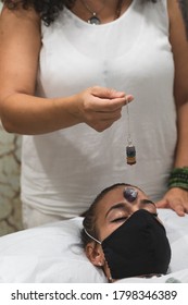 Hand Of A Woman Healer Applying Reiki And Energy Therapies Through The Use Of A Pendulum, To A Young Woman With A Medical Mask During The Coronavirus Crisis