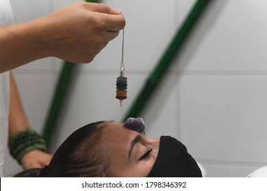 Hand Of A Woman Healer Applying Reiki And Energy Therapies Through The Use Of A Pendulum, To A Young Woman With A Medical Mask During The Coronavirus Crisis