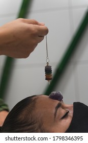 Hand Of A Woman Healer Applying Reiki And Energy Therapies Through The Use Of A Pendulum, To A Young Woman With A Medical Mask During The Coronavirus Crisis