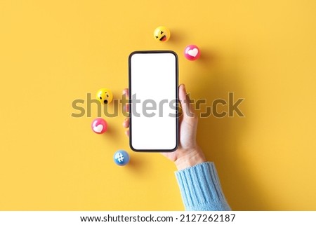 Hand of woman holding blank mobile phone with love, like, comment, hashtag button on yellow background. Social media marketing concept