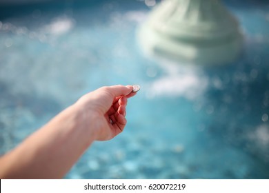 The hand of a woman is holding a coin, about to throw it in the water of a wishing fountain.
