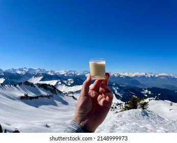 Hand Of Woman Holding Glass Of Eggnog Against Spectacular Winter Panorama In The Austrian Alps. Vorarlberg, Austria. . High Quality Photo