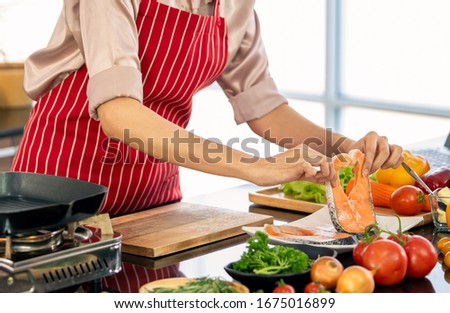 Hand of woman is holding a piece of salmon on a white plate in the kitchen.