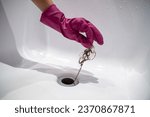 Hand of woman in pink rubber glove taking of hair clump from water drain, blocked pipe in bath. Woman cleaning sewer trap. Bath plug hole blocked with dirt. Problem with shower sewerage in bathroom.