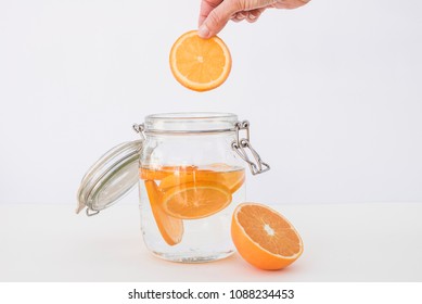 Hand Of Woman Put The Orange Into The Water Glass Jar On White Background