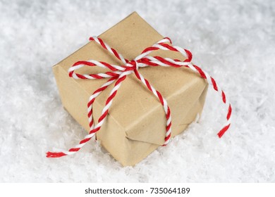 Hand wrapped plain paper small gift box tied with red and white twine, sitting on snow. Christmas, holiday giving or event sale concept.