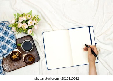 hand write on empty agenda book or notebook and cup of hot coffee with macarons, flowers pot and pillow on white bed