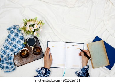 hand write on empty agenda book or notebook and cup of hot coffee with macarons, flowers pot and pillow on white bed
