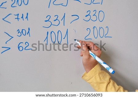A child’s hand writes large math numbers on a whiteboard with copy space, practicing comparisons and early numeracy. Concept of foundational math skills and structured learning.