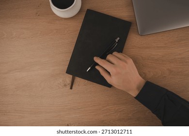 A hand writes notes in a notebook at a home workspace. A cup of coffee sits nearby, next to a laptop on a wooden surface, creating a focused atmosphere for productivity