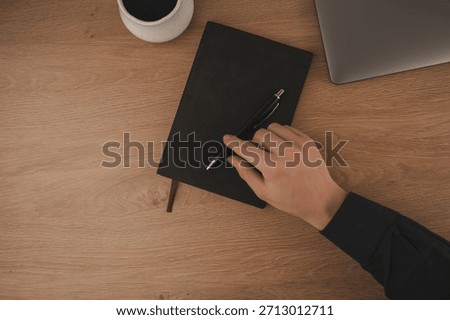 A hand writes notes in a notebook at a home workspace. A cup of coffee sits nearby, next to a laptop on a wooden surface, creating a focused atmosphere for productivity