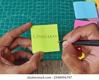 Hand Writing 'Consistent' on Green Sticky Note with Pen on Cutting Mat. - Powered by Shutterstock