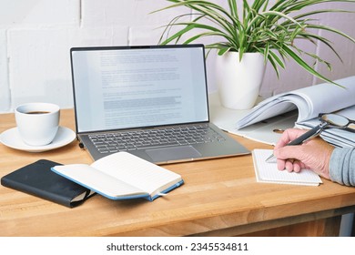Hand writing notes on a wooden desk with laptop, notebooks, file folder and coffee cup, office workplace at home, copy space, selected focus, narrow depth of field