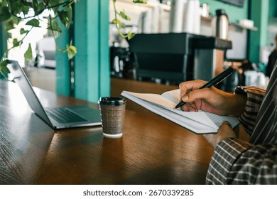 Hand writing notes in open notebook with black pen, laptop and coffee cup on wooden table in modern cafe.
