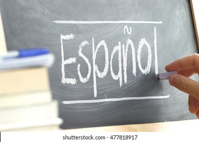 Hand writing on a blackboard in a language class with the word "Spanish" written on it. Some books and school materials.