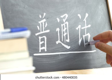 Hand writing on a blackboard in a language class with the text "Chinese Mandarin" written on it. Some books and school materials.