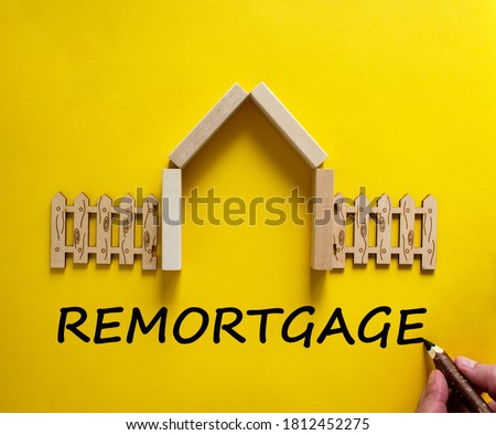 Hand writing 'remortgage' on beautiful yellow background. Model of a wooden house. Male hand, wooden fence. Copy space. Business concept.