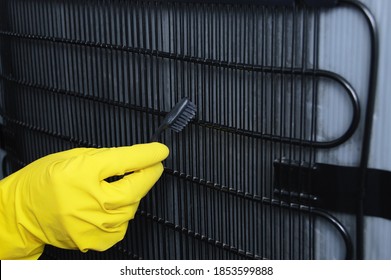 A hand in a yellow glove cleans the fridge radiator grill with a toothbrush close-up.