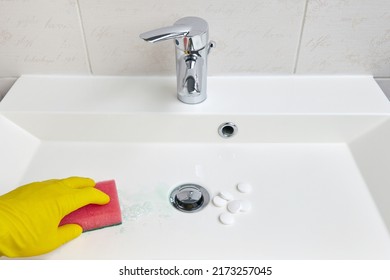 A Hand In Yellow Protective Gloves Cleans The Sink In The Bathroom With A Sponge And A Tablet Bleach Cleaner For Sanitary Equipment In The Bathroom. Cleaning And Cleaning The Washbasin